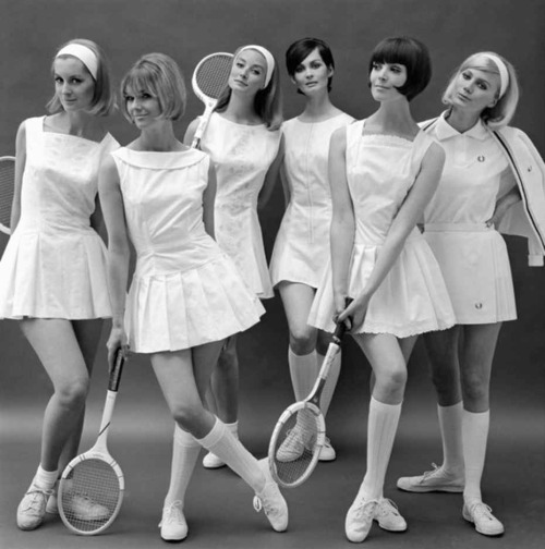 Zes vrouwen in witte tennisoutfits met rackets in de hand poseren samen tegen een effen achtergrond op een zwart-wit foto.