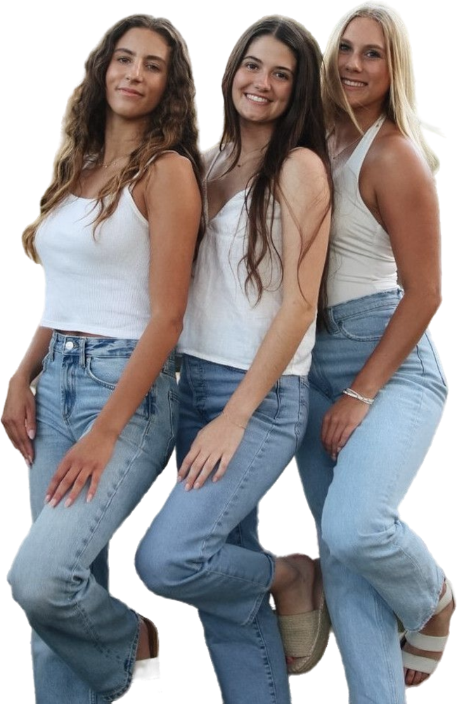 Drie meiden staan lachend tegenover elkaar, elk in een blauwe spijkerbroek en wit topje, poserend met één been iets omhoog.