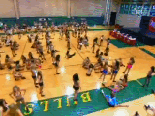 Een grote groep cheerleaders oefent en strekt zich uit op de vloer van een gymzaal, twee cheerleaders worden aangesproken en moeten zich melden in het kantoor.